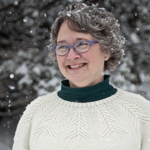 May include: A woman with gray hair wearing a white knitted sweater with a green turtleneck. She is smiling and wearing glasses. Snow is falling around her.