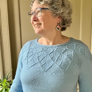 May include: A light blue knit sweater with a geometric diamond pattern across the yoke. The sweater has a crew neck and long sleeves. The person is wearing square earrings with red, blue, and yellow stripes.