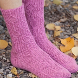 May include: A pair of pink knitted socks with a diamond pattern. The socks are worn on a person's feet and are resting on a bed of small stones and fallen leaves.