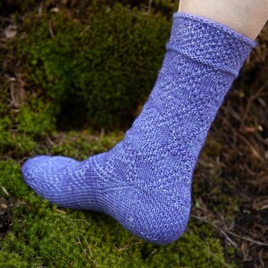 May include: A close-up of a purple knitted sock with a textured pattern. The sock is worn on a foot and is resting on a bed of green moss.