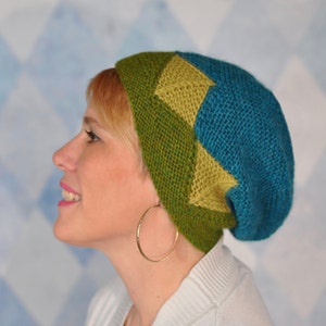 May include: A woman wearing a teal and green knitted hat with a geometric pattern. The hat has a slouchy fit and is made with a soft yarn.