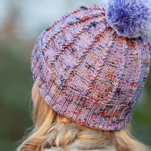 May include: A purple and pink knitted hat with a cable knit pattern and a fluffy purple pom pom on top.