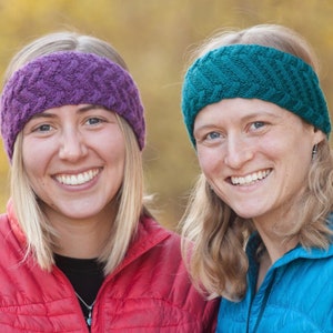 May include: Two women are smiling and wearing knitted headbands. The woman on the left is wearing a purple headband and a red jacket. The woman on the right is wearing a teal headband and a blue jacket.