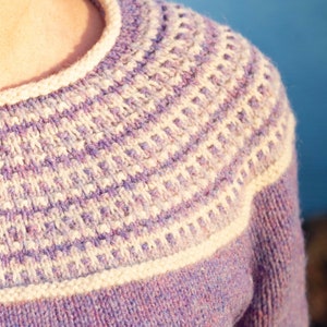 May include: A close-up of a purple and white knitted sweater with a detailed pattern around the neckline.