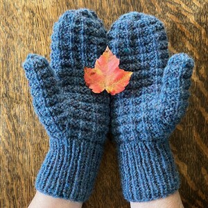 May include: A pair of blue knitted mittens with a textured pattern. A colorful orange and red leaf rests between the mittens. The mittens are made of a soft, warm yarn, perfect for cold weather.
