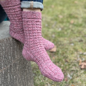 May include: A pair of pink knitted socks with a textured pattern. The socks are being worn on a person's feet and are resting on a tree stump.
