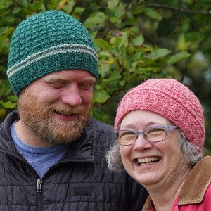 May include: A man and a woman wearing knitted hats. The man wears a green hat with a gray stripe. The woman wears a pink hat. Both are smiling. The background is blurred green foliage.