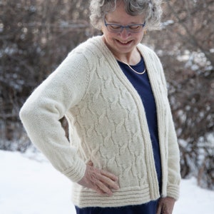 May include: A white knitted cardigan with a cable knit pattern. The cardigan has a V-neck and long sleeves. The person wearing the cardigan is smiling.