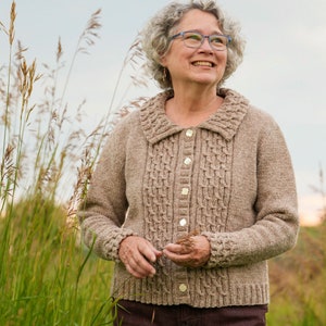 May include: A woman wearing a brown cable knit cardigan with a button-up front and a collared neckline. She is standing in a field of tall grass.