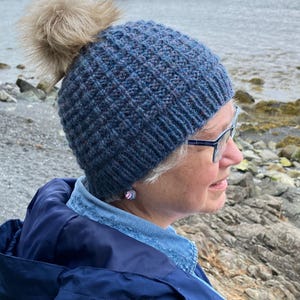 May include: A blue knit hat with a textured pattern and a fluffy, light brown pom-pom. The hat is worn by a person wearing glasses and a blue jacket. The background shows a rocky shoreline and the ocean.