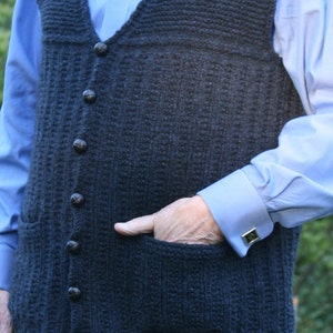 May include: A dark blue knitted vest with a button-up front and a pocket on the left side. The vest has a textured knit pattern and is worn over a light blue button-up shirt.