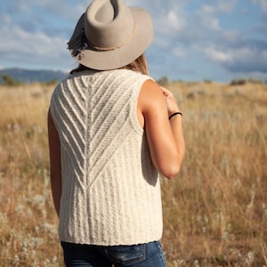 May include: Cream-colored knitted vest with a chevron design on the back. The sleeveless top is worn with blue jeans and a gray hat with a decorative band. The background is a field of dry grass under a blue sky.