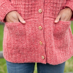 May include: A pink knitted cardigan with a button-up front and two pockets. The cardigan has a textured knit pattern and is decorated with small flower-shaped buttons.