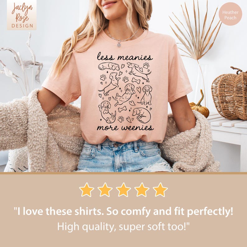 Op de afbeelding: Perzikkleurig T-shirt met de tekst "less meanies more weenies" en een zwarte lijntekening van teckels. De merknaam "Jaclyn Rose Design" staat linksboven. Het shirt is perzikkleurig.