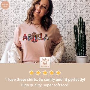 May include: A pink t-shirt with the word "ABUELA" printed on it in colorful letters. The letters are decorated with images of religious figures. The shirt is being worn by a person with a smiling face. The text "Jaclyn Rose DESIGN" is printed below the shirt. Five stars are displayed below the text. The text "I love these shirts. So comfy and fit perfectly! High quality, super soft too!" is printed below the stars.