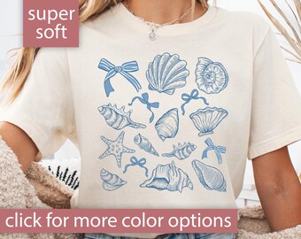 May include: Cream-colored t-shirt featuring blue seashell illustrations and bows. The words "super soft" are in a pink box in the upper left corner. The phrase "click for more color options" is at the bottom.
