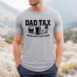 K&ouml;nnte beinhalten: Ein graues T-Shirt mit dem Text "DAD TAX" in Schwarz, das eine Grafik mit Pommes, einem Soda, Keksen und einem Eisbecher zeigt. Der Text unter der Grafik lautet "MAKING SURE IT'S NOT POISON".