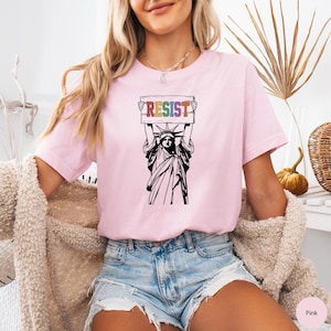 May include: Pink t-shirt with a graphic of the Statue of Liberty holding a sign that says "RESIST" in rainbow colors.