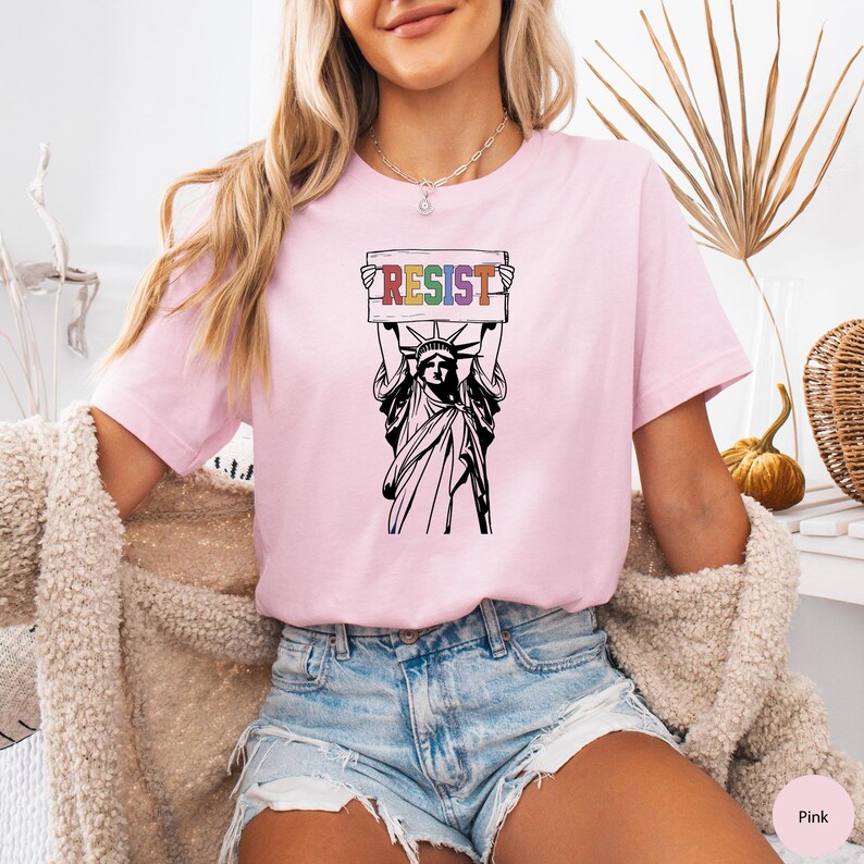 May include: Pink t-shirt with a graphic of the Statue of Liberty holding a sign that says "RESIST" in rainbow colors.