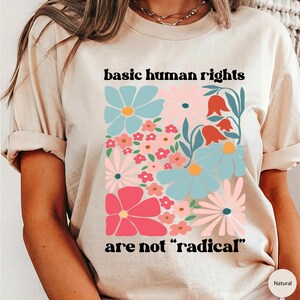 May include: A beige t-shirt with a floral design and the text "basic human rights are not "radical""