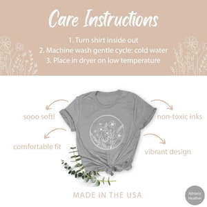 May include: A gray t-shirt with a white floral design in a circle. The shirt is made in the USA and features non-toxic inks. The text "sooo soft!" and "comfortable fit" are written on the left side of the image. The text "non-toxic inks" and "vibrant design" are written on the right side of the image. The text "Care Instructions" is at the top of the image with instructions for washing and drying the shirt. The text "MADE IN THE USA" is at the bottom of the image.