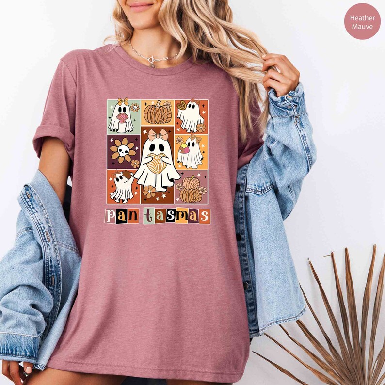 K&ouml;nnte beinhalten: Ein T-Shirt in der Farbe Heather Mauve mit einem Grafikdruck eines Quadrats, das mit niedlichen Geistern, K&uuml;rbissen und Blumen gef&uuml;llt ist. Der Text "Pantasm&aacute;s" ist unter dem Grafikdruck aufgedruckt.