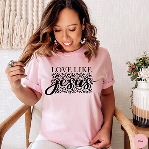 Pode incluir: Camiseta rosa com texto preto que diz "Love Like Jesus" com um design de estampa de leopardo atr&aacute;s da palavra "Jesus".