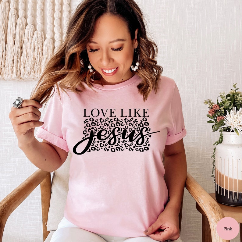 Pode incluir: Camiseta rosa com texto preto que diz "Love Like Jesus" com um design de estampa de leopardo atr&aacute;s da palavra "Jesus".