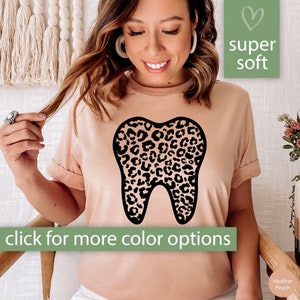May include: A light pink t-shirt with a black tooth graphic featuring a leopard print pattern. The text "super soft" is in a heart-shaped box above the graphic. The text "click for more color options" is below the graphic. The text "Heather Peach" is in the bottom right corner.