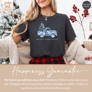 May include: A dark grey heather t-shirt with a blue and white illustration of a pickup truck carrying a Christmas tree. The text "Merry Christmas" is printed below the truck. The shirt is modeled on a person wearing a red and black plaid skirt.