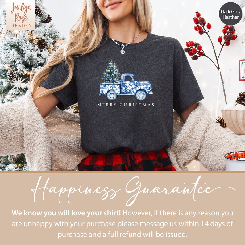 May include: A dark grey heather t-shirt with a blue and white illustration of a pickup truck carrying a Christmas tree. The text "Merry Christmas" is printed below the truck. The shirt is modeled on a person wearing a red and black plaid skirt.