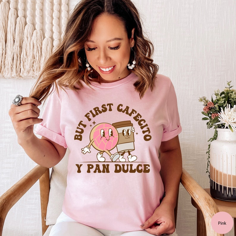 以下が含まれることがあります： 「But first cafecito y pan dulce」というテキストと、ドーナツとコーヒーカップのグラフィックが描かれたピンクのTシャツ。