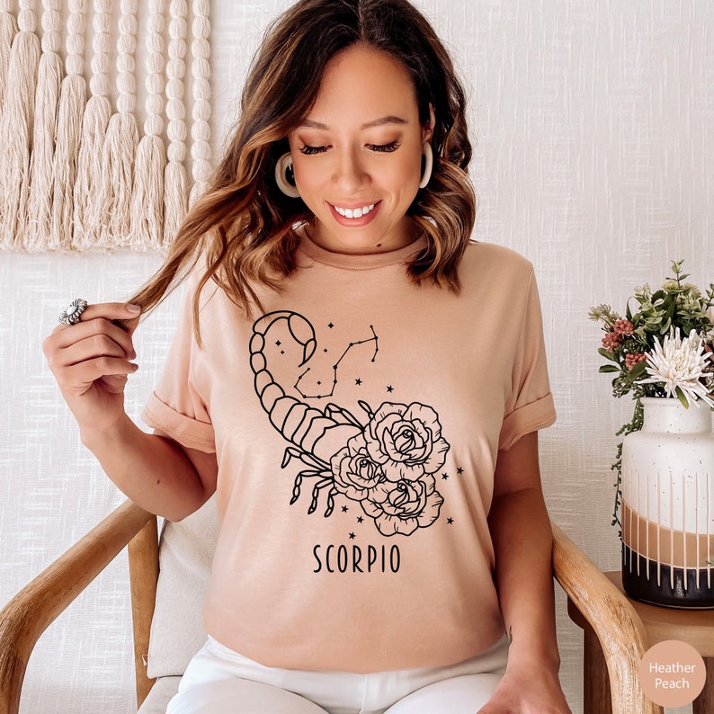 Pu&ograve; includere: Una maglietta color pesca chiaro con un disegno a linea nera di uno scorpione con rose e stelle. Il testo "SCORPIO" &egrave; stampato sotto lo scorpione.