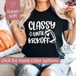 May include: Black t-shirt with white text that reads "Classy Until Kickoff" and a white football with a heart on it.