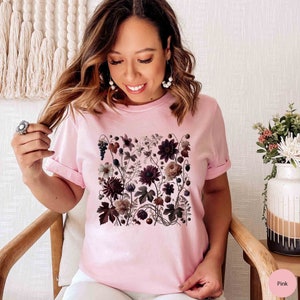 Op de afbeelding: Een roze T-shirt met een bloemenprint met donkerroze, witte en bruine bloemen. De bloemen zijn in een vierkant patroon op de voorkant van het shirt gerangschikt.
