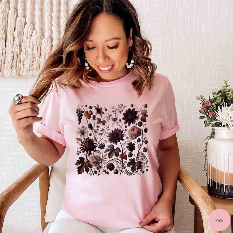 Op de afbeelding: Een roze T-shirt met een bloemenprint met donkerroze, witte en bruine bloemen. De bloemen zijn in een vierkant patroon op de voorkant van het shirt gerangschikt.