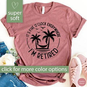 May include: Mauve-colored t-shirt with the text "It's Five O'Clock Everywhere, I'm Retired" in a black circular design featuring palm trees and a hammock. The shirt also has the words "super soft" and "click for more color options".