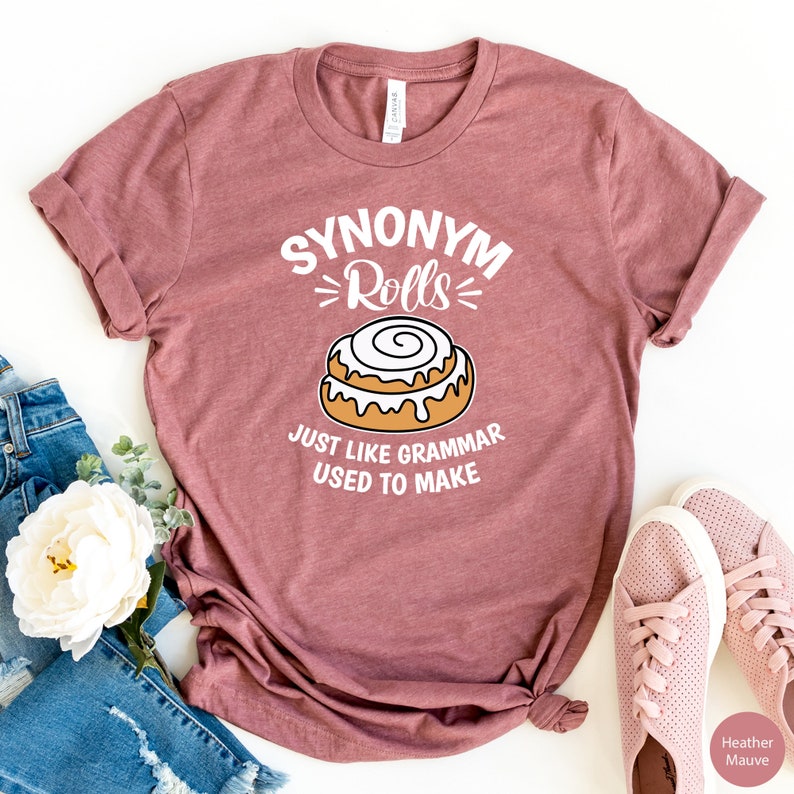 May include: A heather mauve t-shirt with the text "Synonym Rolls" and a cartoon cinnamon roll. The text below the image says "Just like grammar used to make".