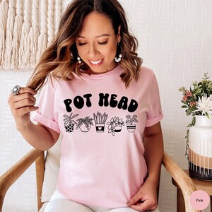 May include: A pink t-shirt with the text "POT HEAD" in black, featuring a row of five potted plants with different designs.