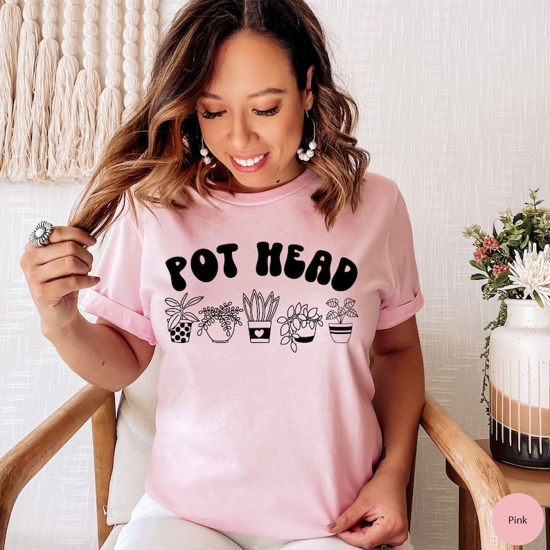 May include: A pink t-shirt with the text "POT HEAD" in black, featuring a row of five potted plants with different designs.