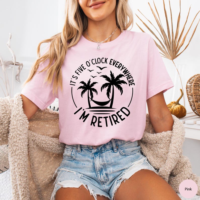 May include: Pink t-shirt with the text "It's Five O'Clock Everywhere, I'm Retired" in a black circular design featuring palm trees, a hammock, and birds. The shirt is worn with denim shorts.