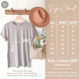 Op de afbeelding: Een lichtgrijs unisex jersey t-shirt hangt aan een houten rek. Er wordt een maattabel weergegeven, met afmetingen in inches. Een bruine hoed en een beige tas met bloemen staan ook op de afbeelding. Het shirt is gemaakt van 100% katoen.