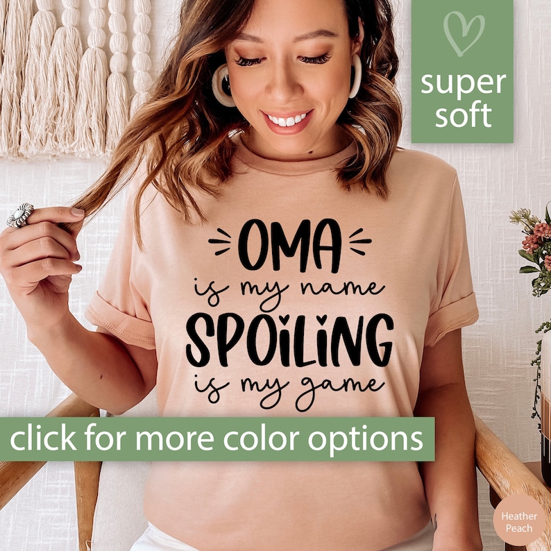 May include: A pink t-shirt with black text that reads "OMA is my name SPOILING is my game". The shirt is super soft and has a heart design.