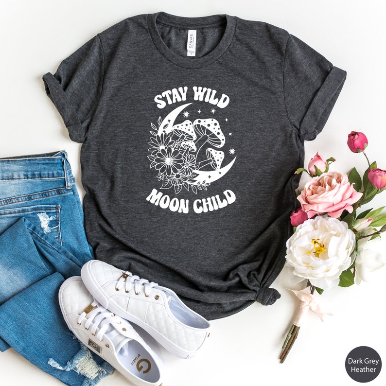 Puede incluir: Camiseta de color gris oscuro jaspeado con un gr&aacute;fico blanco de una luna creciente con hongos y flores a su alrededor. El texto "Stay Wild Moon Child" est&aacute; impreso en la camiseta.