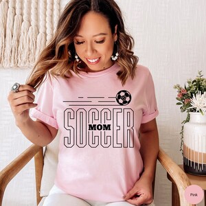 Pu&ograve; includere: Maglietta rosa con una grafica di un pallone da calcio nero e la scritta "Soccer Mom" stampata su di essa.