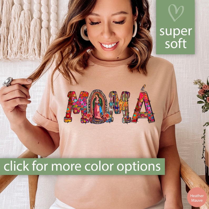 May include: A light pink t-shirt with the word "MOM&Aacute;" in colorful, patterned letters. The letters are decorated with flowers, geometric shapes, and a small image of the Virgin Mary. The shirt is described as "super soft" and has a "click for more color options" call to action.