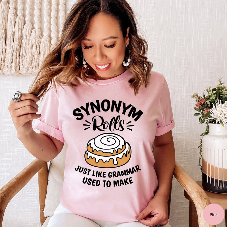 May include: Pink t-shirt with the text "Synonym Rolls" and a cinnamon roll graphic. The text below the graphic reads "Just like grammar used to make."