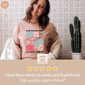 May include: A pink t-shirt with a floral design and the text "basic human rights are not 'radical'" printed on the front. The shirt is being worn by a person. The text "Jaclyn Rose DESIGN" is printed below the design. The shirt has a five-star rating.