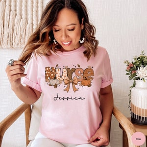 May include: Pink t-shirt with a fall-themed design that says "Nurse Jessica". The design features a bow, pumpkins, and leaves in shades of brown, orange, and white.