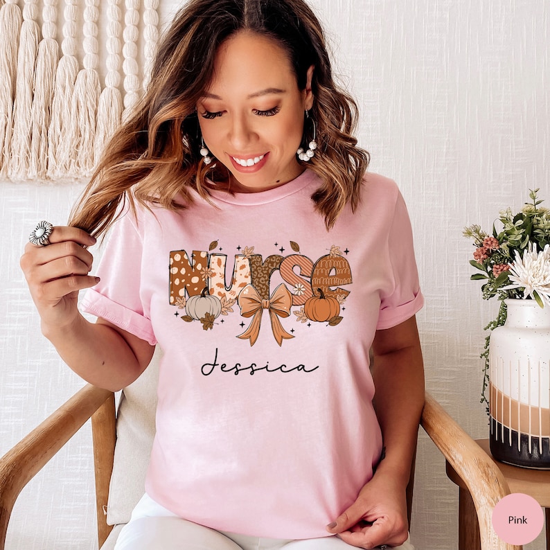 May include: Pink t-shirt with a fall-themed design that says "Nurse Jessica". The design features a bow, pumpkins, and leaves in shades of brown, orange, and white.